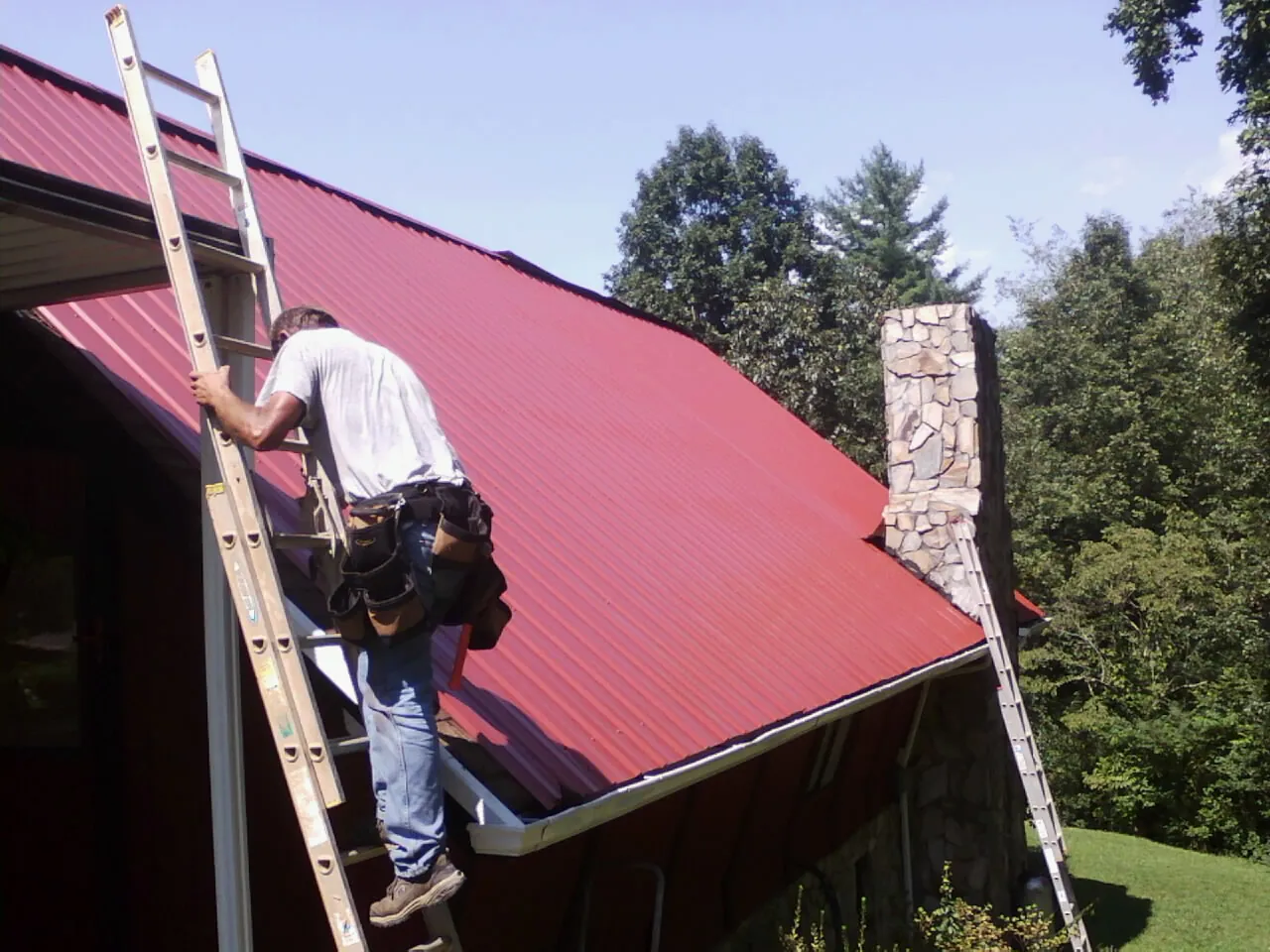 Roofer on a ladder beside a red metal roof and stone chimney — Soffit & Fascia Repair by Roofing Brothers in Solen